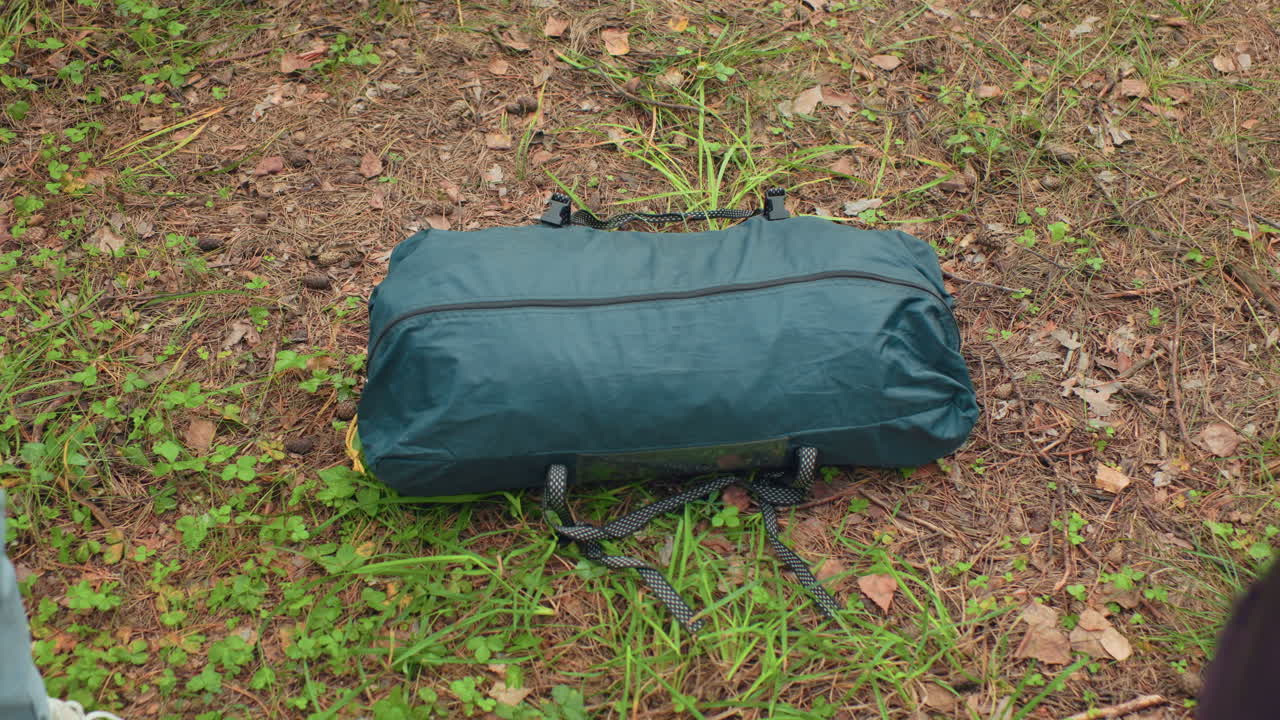 close up view of camper lowering green bag onto forest ground, surrounded by leaves, pine needles, and natural woodland terrain, capturing moment of resting