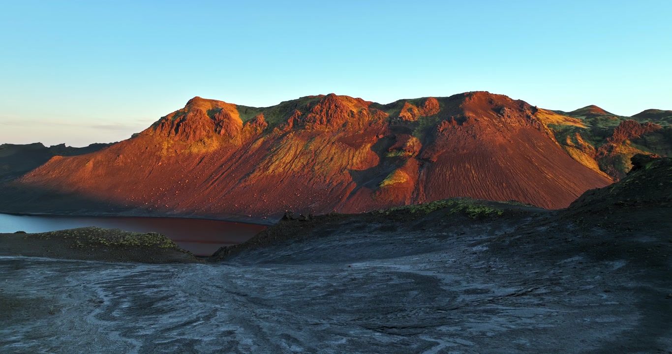 Golden-hour light illuminates rugged volcanic mountains above dark lava plains and a lakeshore, revealing dramatic textures, warm colors, and remote highland wilderness.