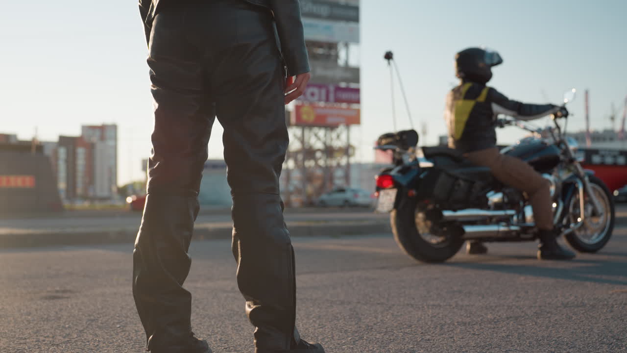 Passenger in leather outfit approaches parked motorcycle preparing to climb behind rider seated on power bike, holding balance as sunlight highlights street and blurred cars in urban background
