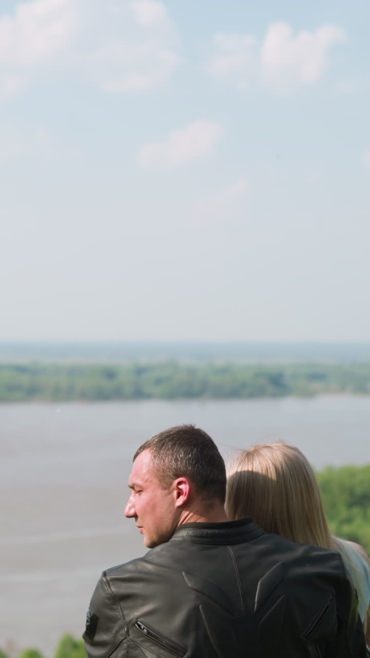 Man in moto suit embraces beloved woman enjoying wonderful view of tranquil river from hill on sunny spring day backside view slow motion