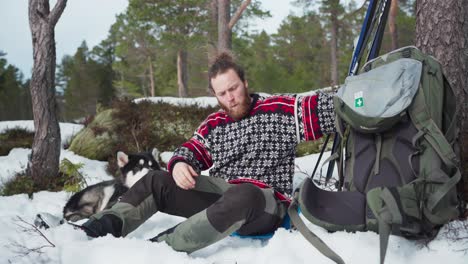 A-Traveler-Man-Eating-And-Resting-With-Alaskan-Malamute-On-A-Winter-Season-At-Indre-Fosen,-Norway