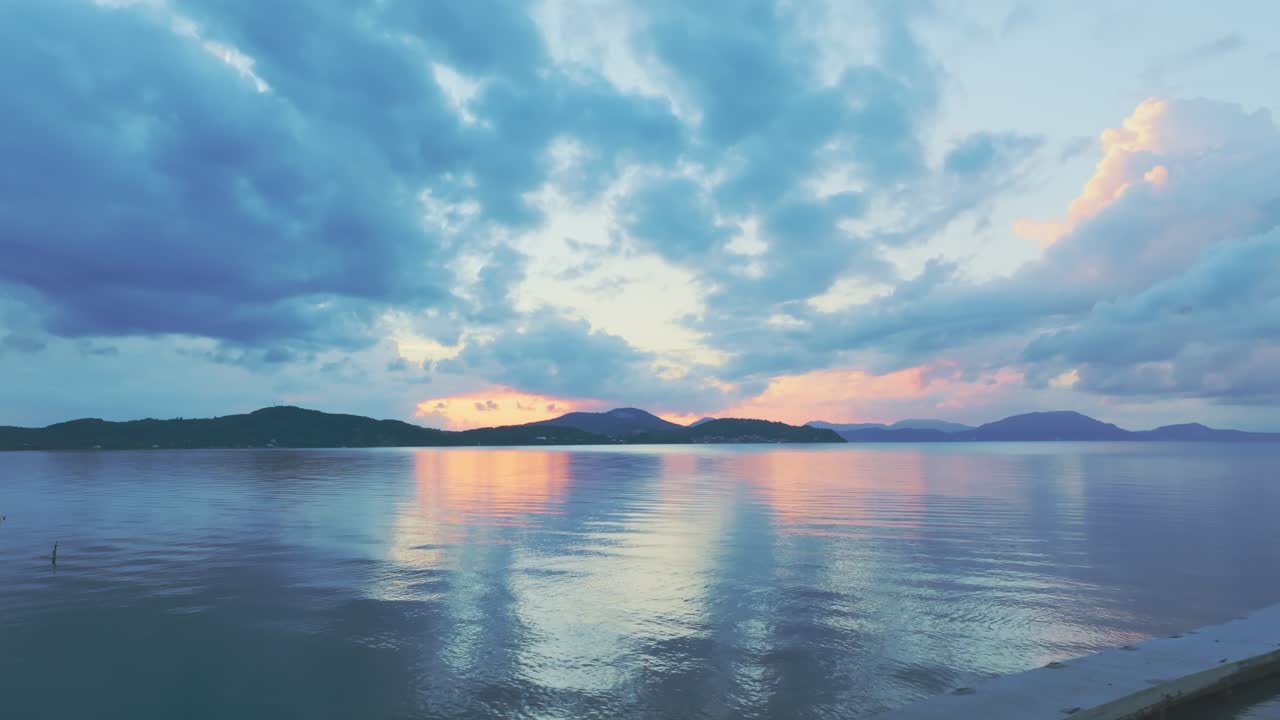 Serene coastal scene at sunset featuring a wooden pier, calm reflective waters, and vivid clouds over distant hills. A peaceful and scenic landscape perfect for travel and nature themes. Greece, Corfu
