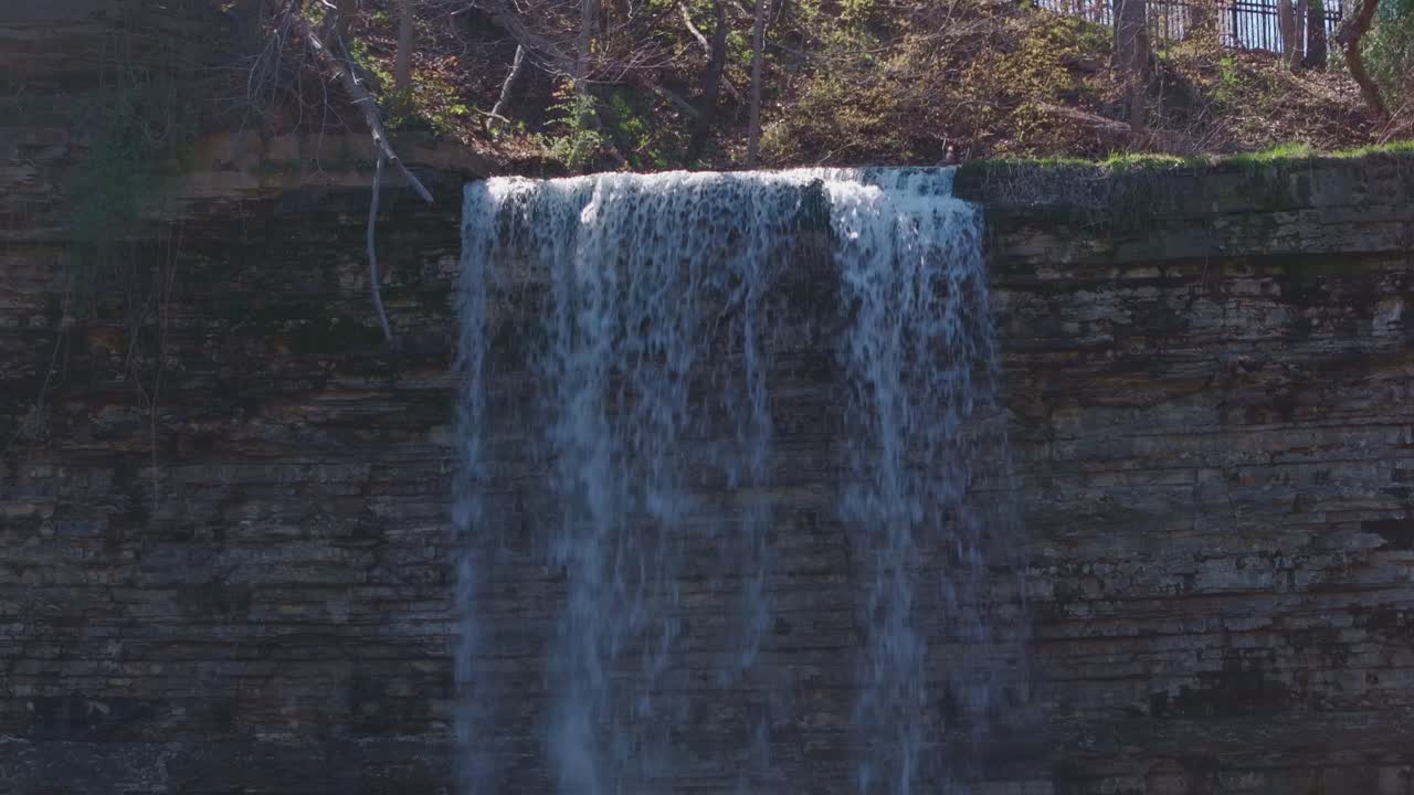 cascada en cascada sobre un acantilado rocoso con vegetación exuberante por encima, filmado en un día soleado