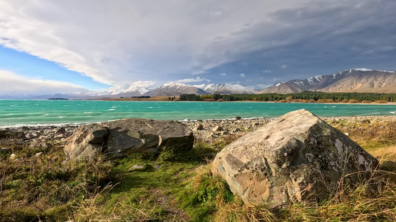 Sweeping panoramic camera movement reveals Lake Tekapo’s turquoise waters, rocky shoreline, alpine grasses, and distant snow-capped mountains beneath dramatic, stormy skies
