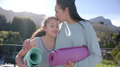 happy biracial mother and daughter standing with yoga mats on terrace in sunny day, slow motion