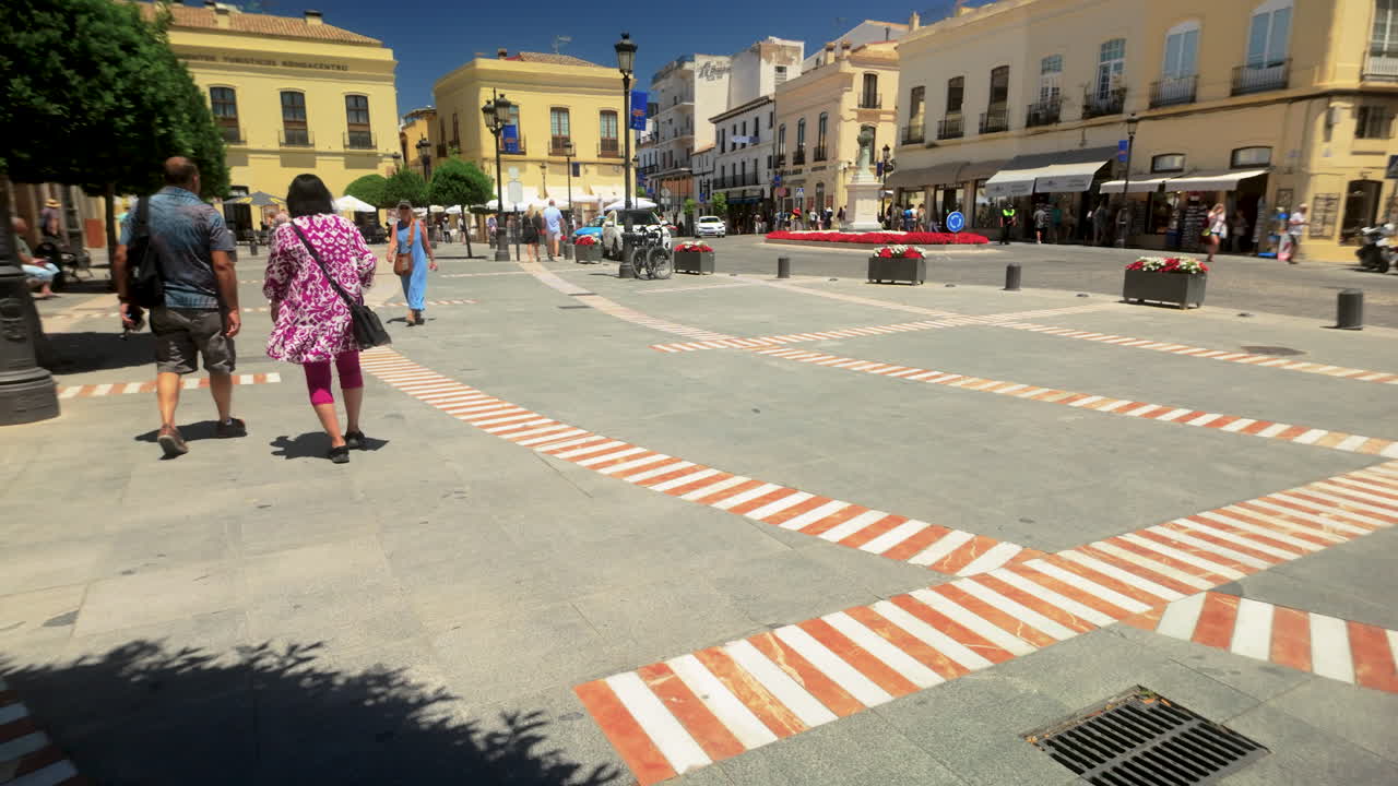 People strolling through a sunny square in Ronda, Spain, surrounded by historic buildings and vibrant trees, showcasing the town’s lively culture