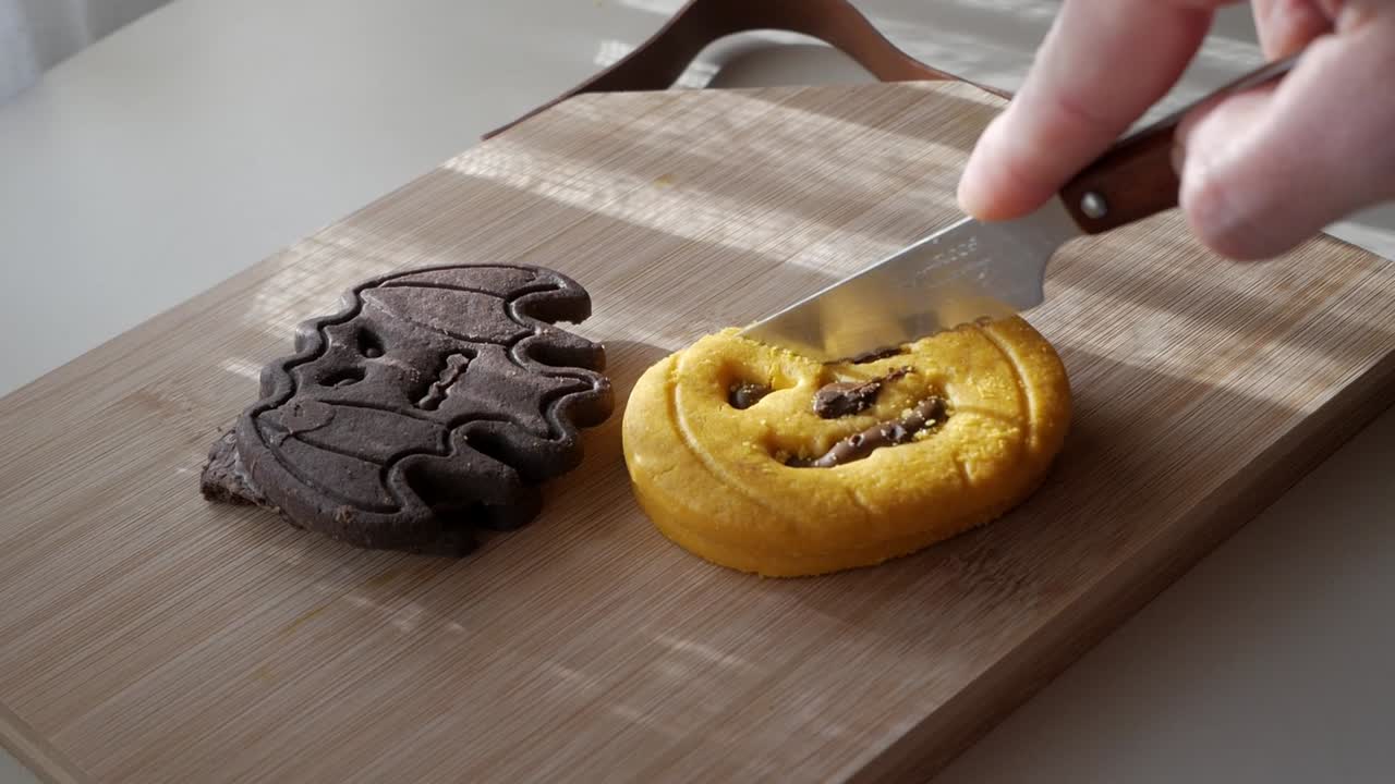 Side shot of person slicing Halloween pumpkin and bat shaped cookies on wooden board under warm sunlight