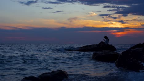 Mädchen,-Das-Die-Ruhe-Des-Schönen-Felsigen-Strandes-Bei-Sonnenuntergang-Mit-Dunklen-Wolken-Am-Bunten-Himmel-Genießt