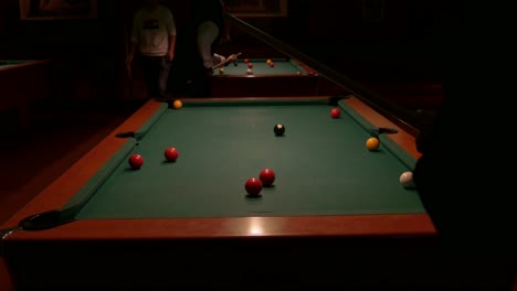 over the shoulder, row of two pool tables and silhouette of person shooting balls with a cue stick