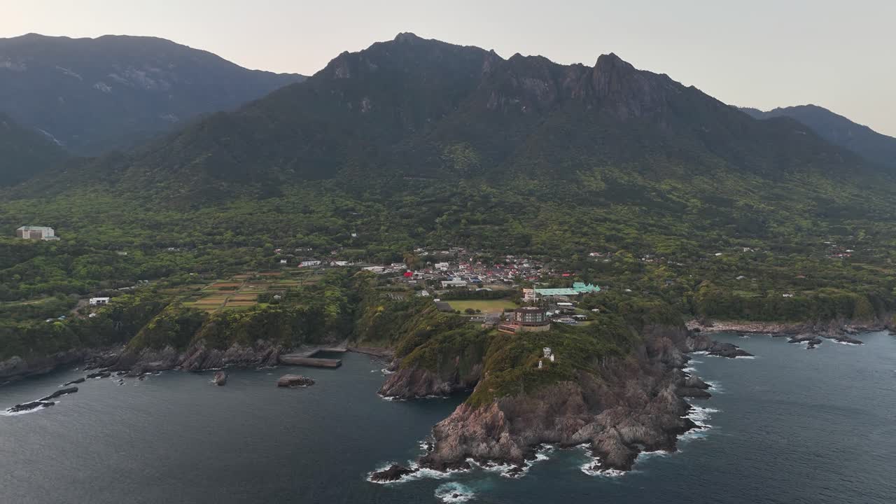 Aerial panorama revealing Onoaida nestled between verdant mount Mocchomu and rocky shoreline, Yakushima Island stretching toward expansive Pacific waters in Japan, drone slowly pulling out
