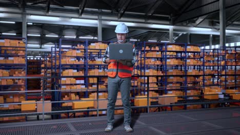 cuerpo lleno de ingeniero masculino asiático con casco de seguridad mirando una computadora portátil y mirando a su alrededor mientras estaba de pie en el almacén con estantes llenos de bienes de entrega