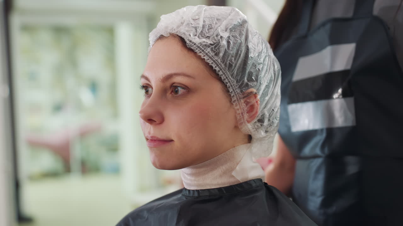 Una mujer blanca con gorro protector en un salón de belleza; una clienta sentada tranquilamente con una redecilla después de la aplicación de un producto mientras la estilista controla el tiempo y la comodidad. El fondo neutro y las herramientas profesionales dan una idea del entorno.