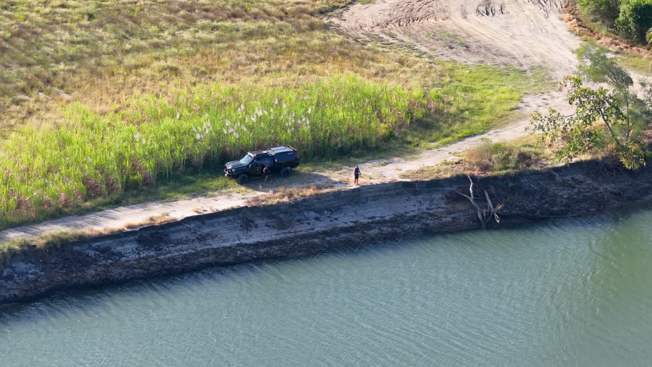 Aerial footage captures a vehicle and person by a riverbank in Port Douglas. Bright daylight enhances the natural setting