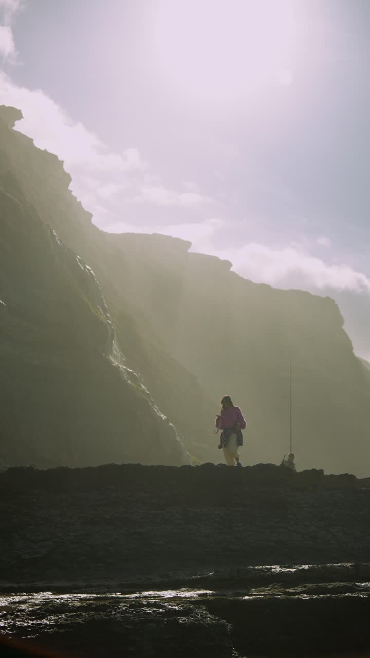 Donna in piedi su costa rocciosa con scogliere retroilluminate e cascata