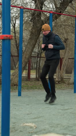 un entrenador corre a toda velocidad en un estadio, rodeado de postes azules y rojos, con un banco cerca, con árboles estériles y coches y personas que pasan visibles en el fondo