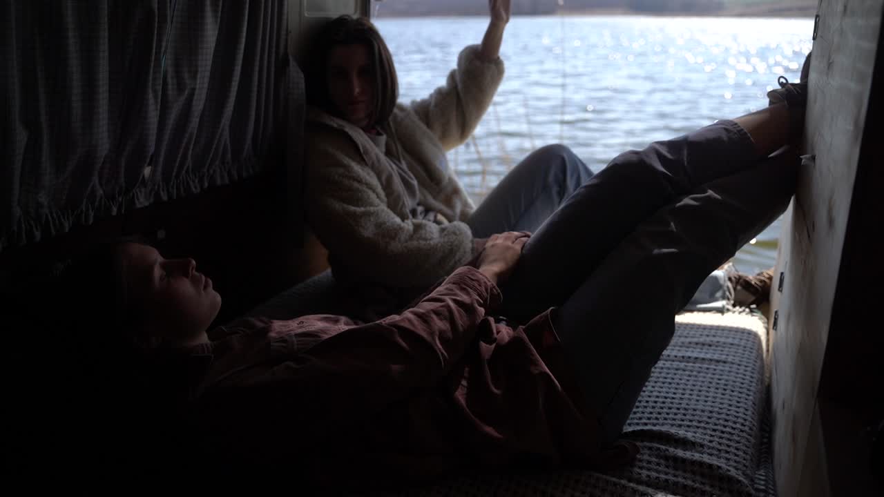 두 소녀가 캐러밴 뒤쪽 호수 옆에 누워 있습니다. 그들은 아름다운 경치를 가지고 있어요.