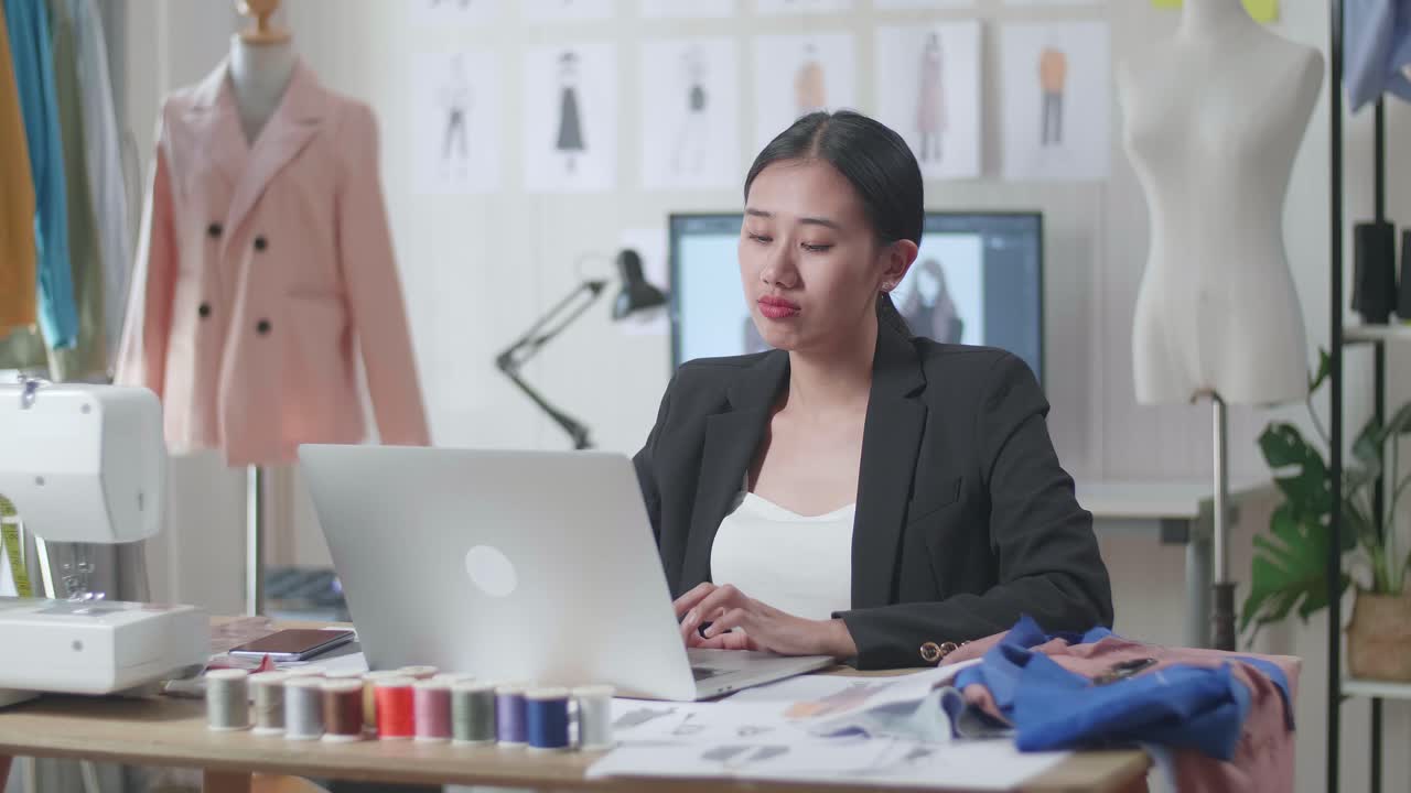 비즈니스 슈트 를 입은 아시아 여성 디자이너 가 노트북 을 사용 하고 스튜디오 에서 옷 을 디자인 하는 동안 허리 통증 을 겪고 있다