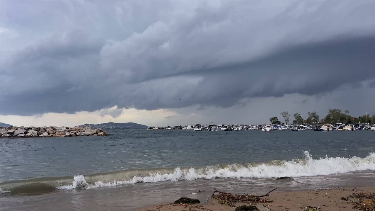 Dramatic dark storm clouds gather over a rough sea. Waves crash on the shore near a stone breakwater and a marina with boats on a gloomy autumn day