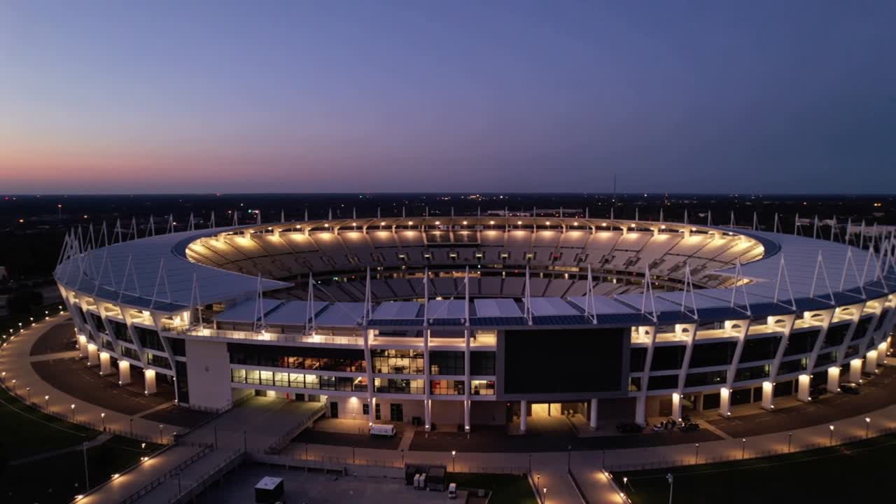 Drone in orbita che riprende uno stadio circolare in periferia al tramonto, mostrando l'anello del tetto illuminato e il display