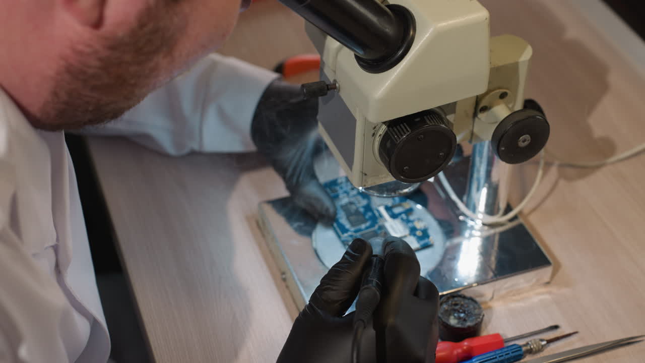 vista hacia abajo de un técnico soldando una placa de circuito bajo un microscopio, el espacio de trabajo incluye varias herramientas y equipos