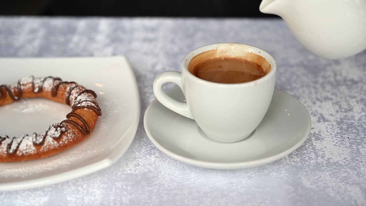 Close up of a man poring cream into his coffee near a pastry on a table
