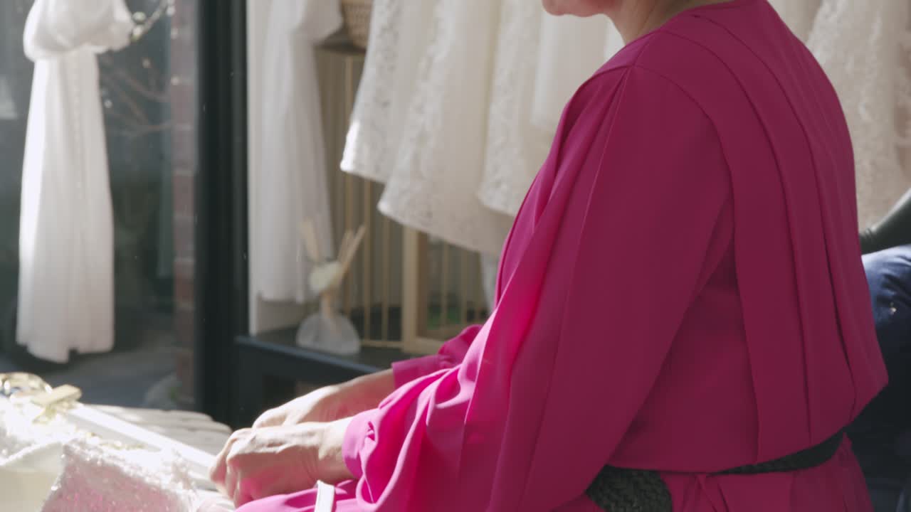 Asian woman in pink dress delicately examines lace fabric in a sunlit bridal boutique, surrounded by elegant dresses and soft shadows, highlighting her meticulous care