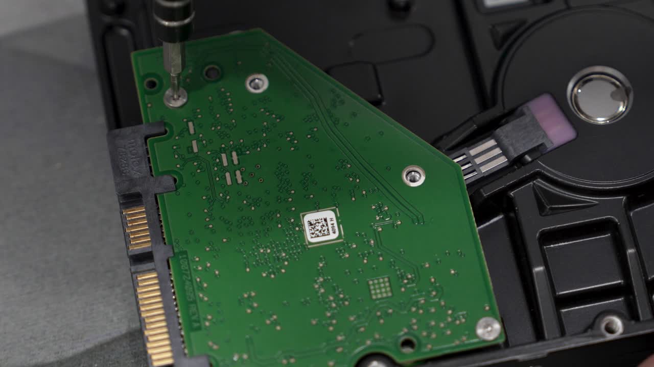 A person is taking apart a hard drive using a screwdriver. They carefully remove screws and handle the parts. Tools and a work surface are visible in the background