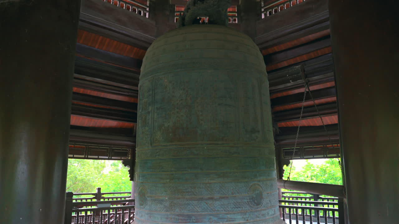 Una toma detallada con inclinación ascendente de la enorme Gran Campana en la Pagoda Bai Dinh, mostrando intrincados grabados de bronce y arquitectura tradicional de madera en el campanario de Ninh Binh, Vietnam