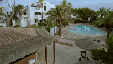 Aerial-view-of-a-tourist-resort-in-Mallorca,-Spain,-featuring-an-accommodation-building-with-a-front-yard-adorned-with-chairs-and-tables-with-an-open-swimming-pool