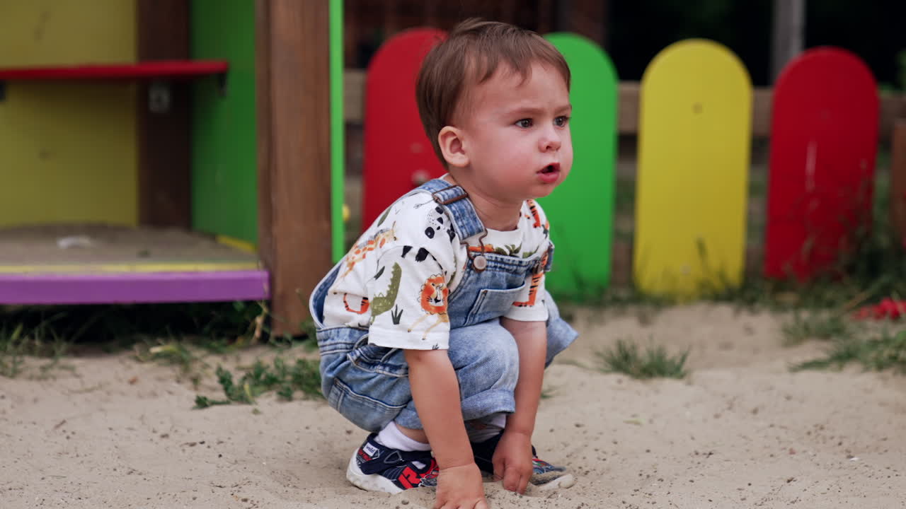 Lovely baby boy sits deepening his hands into the sand. Little kid looks ahead thoughtfully.