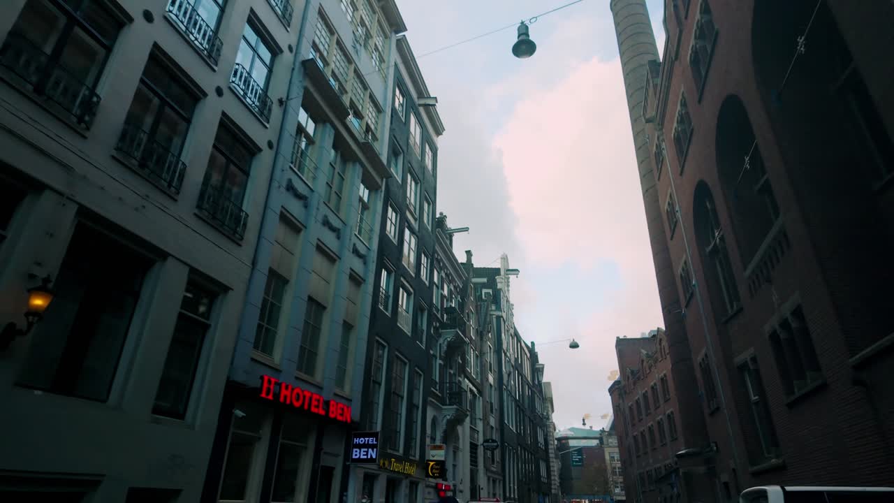 Narrow street in Amsterdam near Damrak with classic Dutch architecture, streetlights, and a view of a tall tower against a partly cloudy sky. Location: Amsterdam, Netherlands