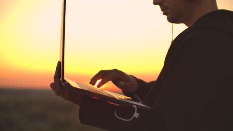 Hacker-using-laptop-on-rooftop-with-city-view-and-forex-chart.-Hacking-and-stats-concept.-A-man-at-sunset-in-slow-motion-writing-software-code-on-a-laptop