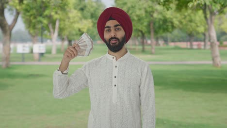 egoistic sikh indian man using money as fan with cunning smile in park