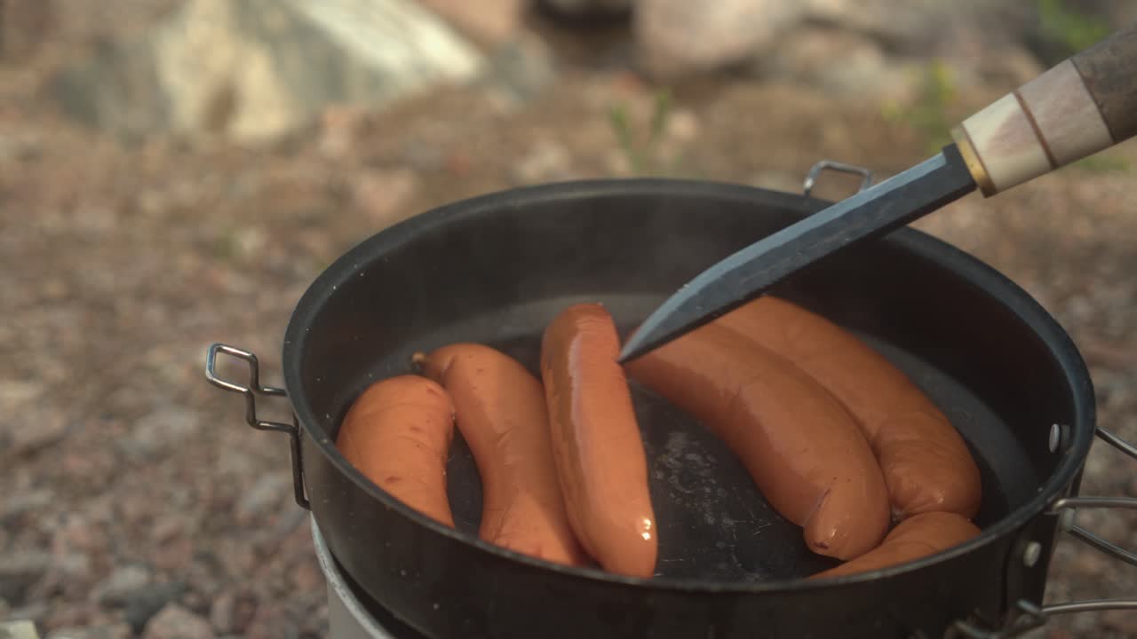 las salchichas se cocinan en una sartén en una estufa al aire libre, empujando con un cuchillo