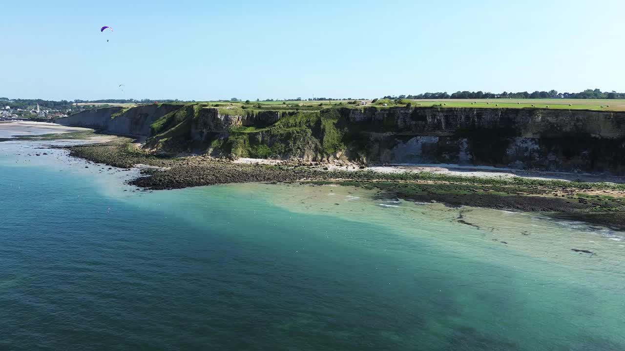 Drone footage gliding along the Arromanches cliffs, showing their majestic drop into the blue sea, framed by lush green fields under a cloudless sky