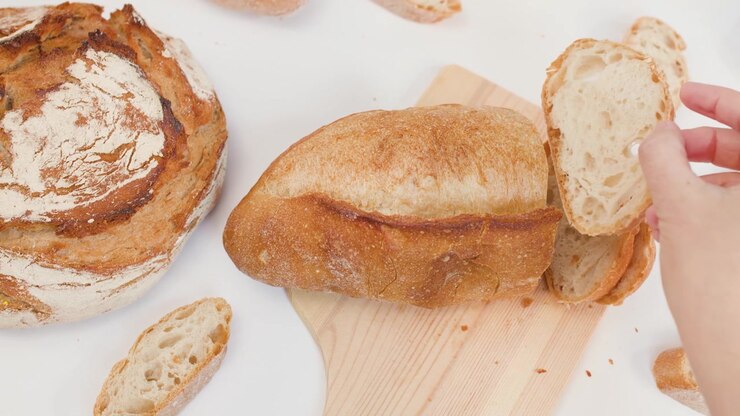 Top-down view of hands slicing crusty sourdough loaf on wooden board with bright lighting.