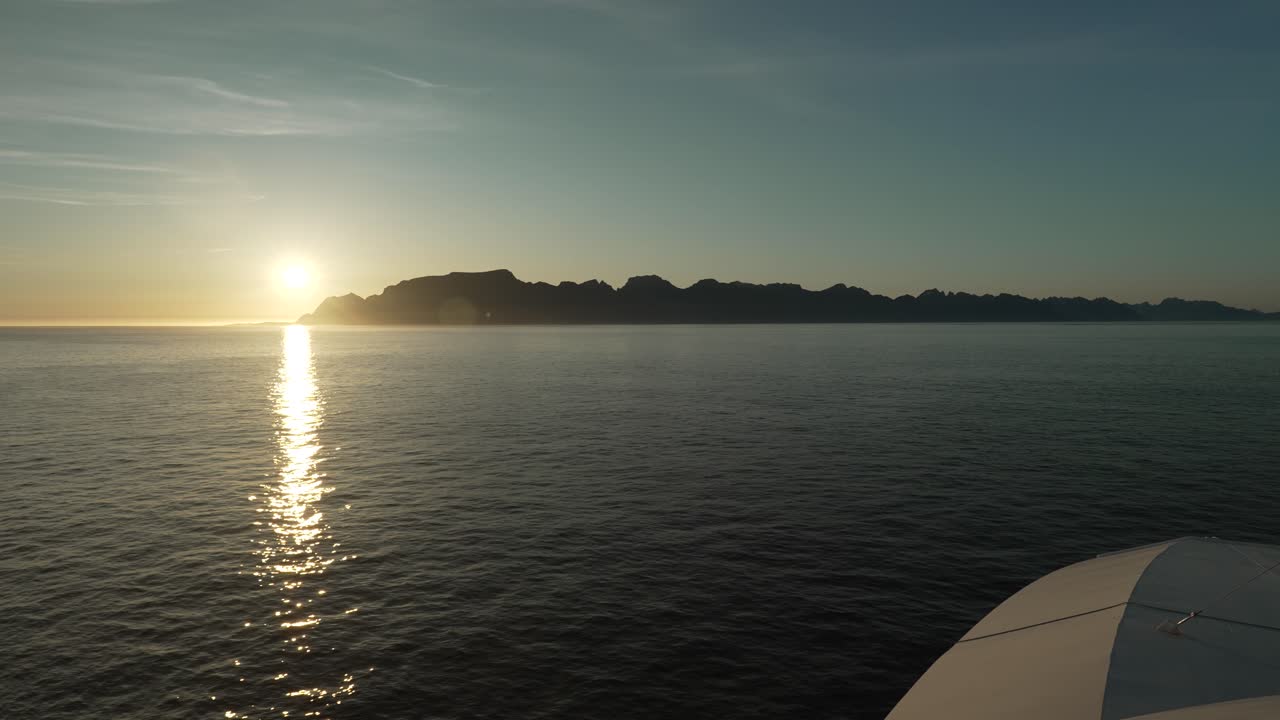 A ferry approaches the Lofoten Islands on a calm summer evening, as the sun sets behind the majestic mountains. Golden light reflects on the sea, creating a breathtaking and serene atmosphere