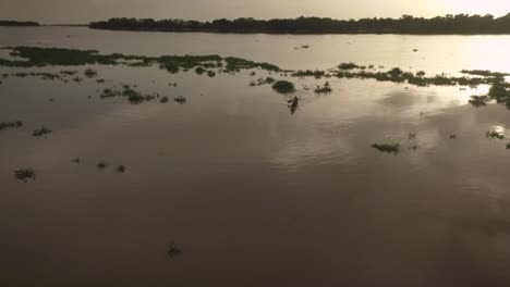 Vista-De-Drones-De-Una-Pequeña-Canoa-Indígena-Cruzando-Un-Montículo-De-Algas-Flotantes-En-El-Río-Orinoco-Durante-La-Puesta-De-Sol