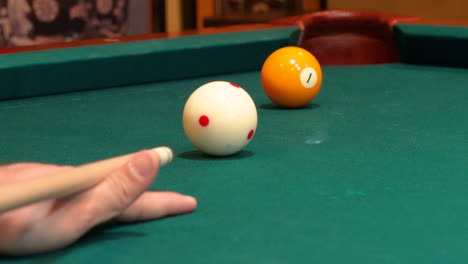 person playing pool shoots yellow solid 1 ball into corner pocket with cue ball drawing or spinning backwards towards camera after stroke, closeup open bridge hand with wooden cue stick and green felt