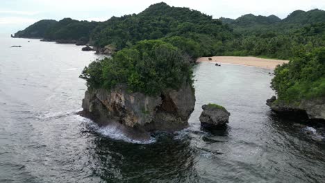 an aerial shot of a massive rock in the ocean, waves crashing into it, with an idyllic sand beach in the background in catanduanes, philippines