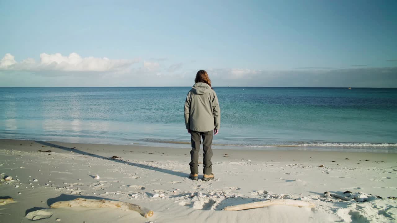 Standing woman in hooded jacket at sandy shoreline, watching distant sailboat as waves lap