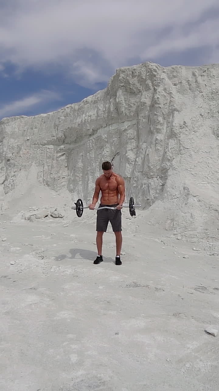 Sporty man lifting heavy barbell in the mountains. Bodybuilder doing his workout on the white background of a rocky hill. Slow motion. Vertical video