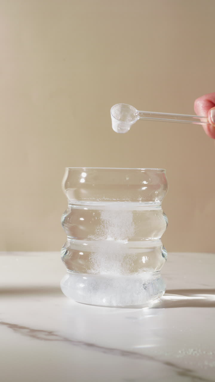 A translucent scoop of fine white powder, possibly creatine, magnesium, or electrolytes, is poured into a clear glass of water, highlighting a clean supplement routine on a marble surface.