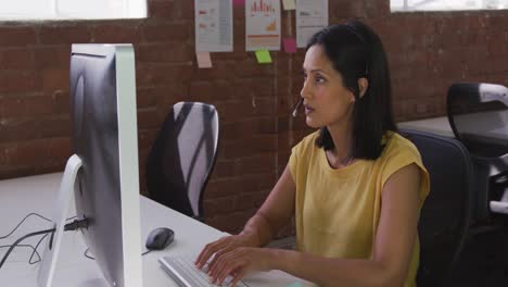 Mixed-race-businesswoman-wearing-headset-sitting-at-desk-using-computer