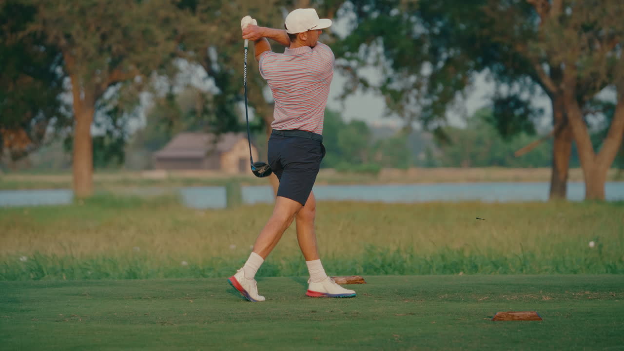 A male golfer takes a powerful swing in slow motion off the tee box. The background features a calm water view and glowing sunset, creating a cinematic moment of focus and energy on the course.