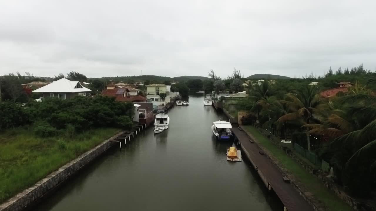 Aerial footage starting from the water, revealing boats docked in a canal, surrounded by lush greenery, palm trees, and large homes in Búzios, Brazil, perfect for real estate and tropical content.