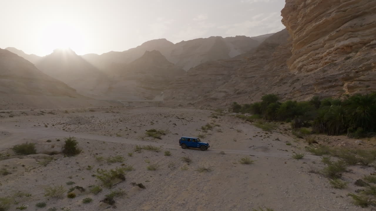 A blue jeep drives through a dry, desert landscape near the coast of Oman