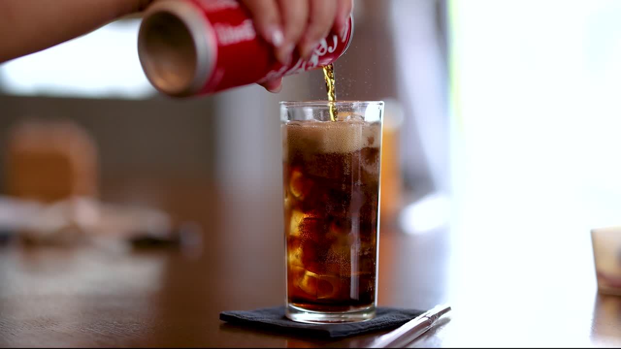 Hand pours cola into tall glass with ice, natural daylight, shallow focus, relaxed dining atmosphere