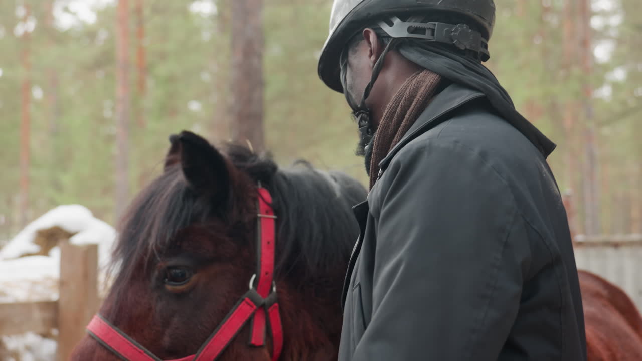 Un hombre negro abraza a un caballo castaño en la nieve; cuidador del establo con casco y bufanda; suave caricia contra el cabestro rojo; valla de madera y bosque de pinos al fondo; primer plano emocional que transmite confianza