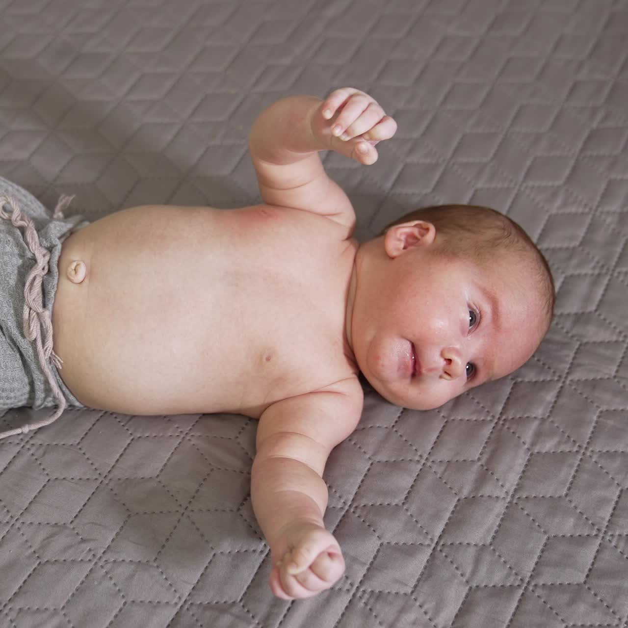 Little boy wearing grey pants lies on the bed. Cute newborn sneezes funnily. View from above on the child with naked belly